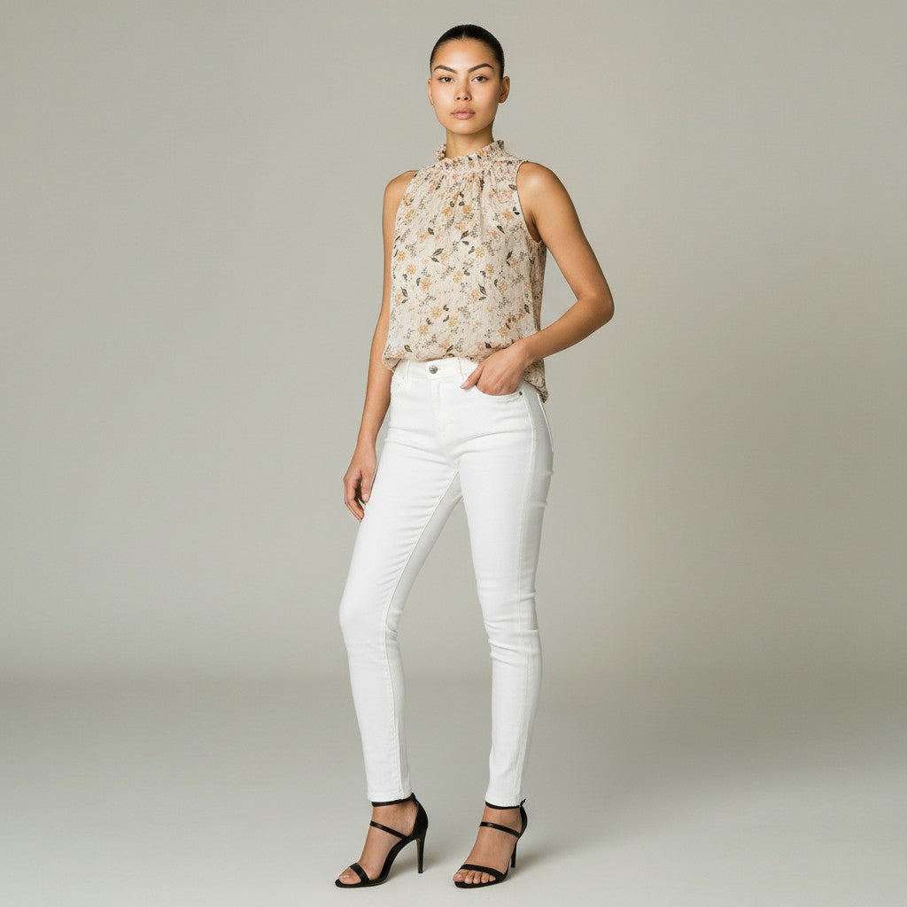 A model stands against a neutral light gray studio background wearing a sleeveless beige and brown floral print top, white skinny jeans, and black strappy high-heeled sandals.