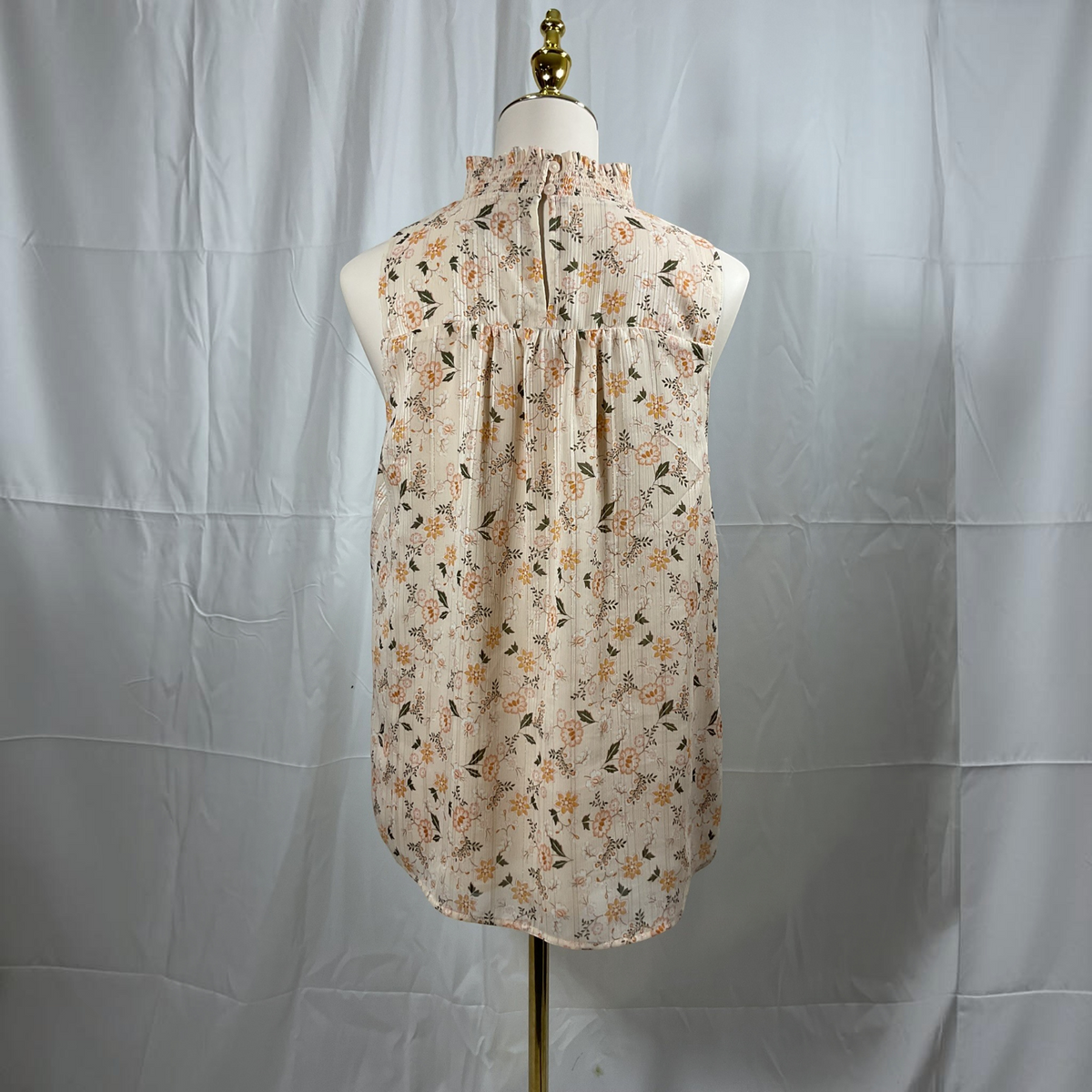 A sleeveless high-neck blouse with a gathered, pleated back, featuring an all-over floral print in orange, yellow, and green on a light beige background. The blouse is displayed on a white dress form with a gold stand against a wrinkled light gray backdrop.