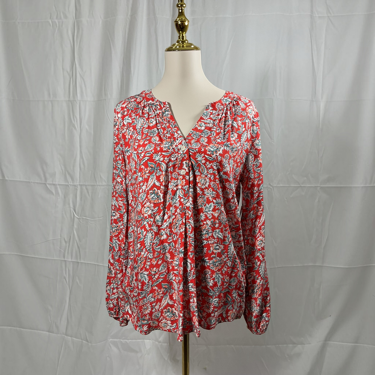 A long-sleeve blouse with a red, white, and gray floral pattern, displayed on a cream mannequin torso with a gold neck cap against a wrinkled white backdrop.
