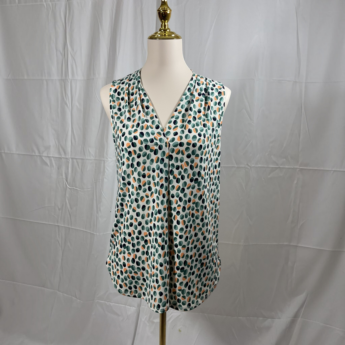 A sleeveless V-neck blouse with a green, black, and orange polka dot pattern, displayed on a white mannequin torso against a wrinkled white fabric backdrop.