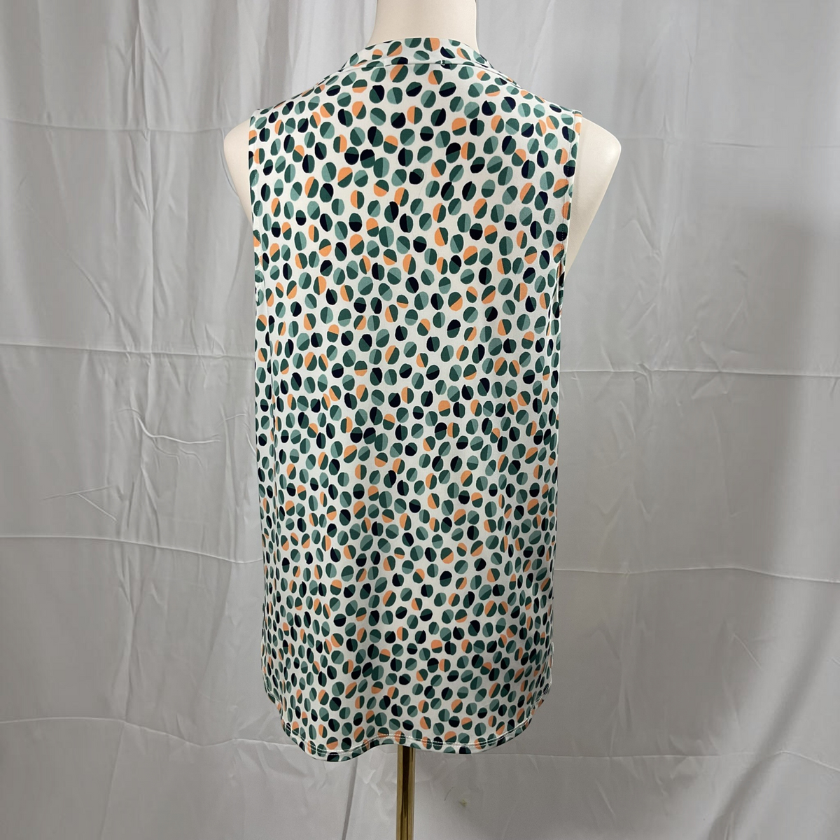 A sleeveless top displayed on a mannequin torso, featuring an abstract spotted pattern in teal, orange, black, and off-white on a light background, set against a wrinkled white backdrop.