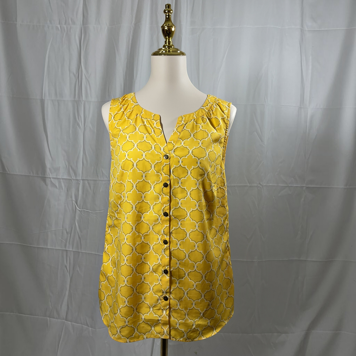 A yellow sleeveless blouse with a white geometric trellis pattern and a button-down front, displayed on a white dress form mannequin against a wrinkled gray fabric backdrop.