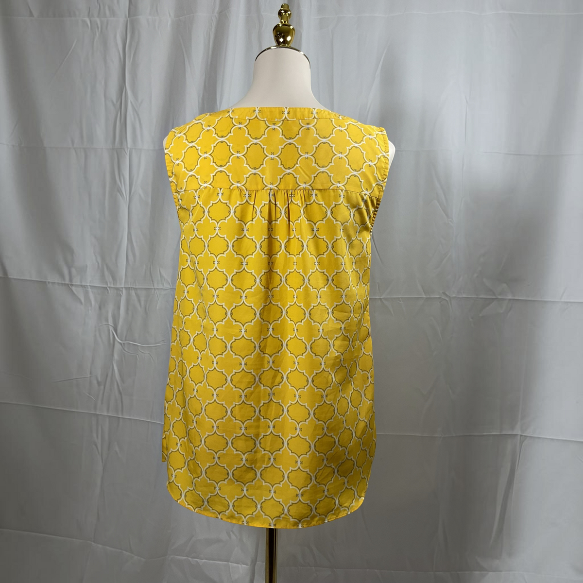 A yellow sleeveless top with a white geometric trellis pattern, displayed on a dress form mannequin against a wrinkled light gray backdrop.