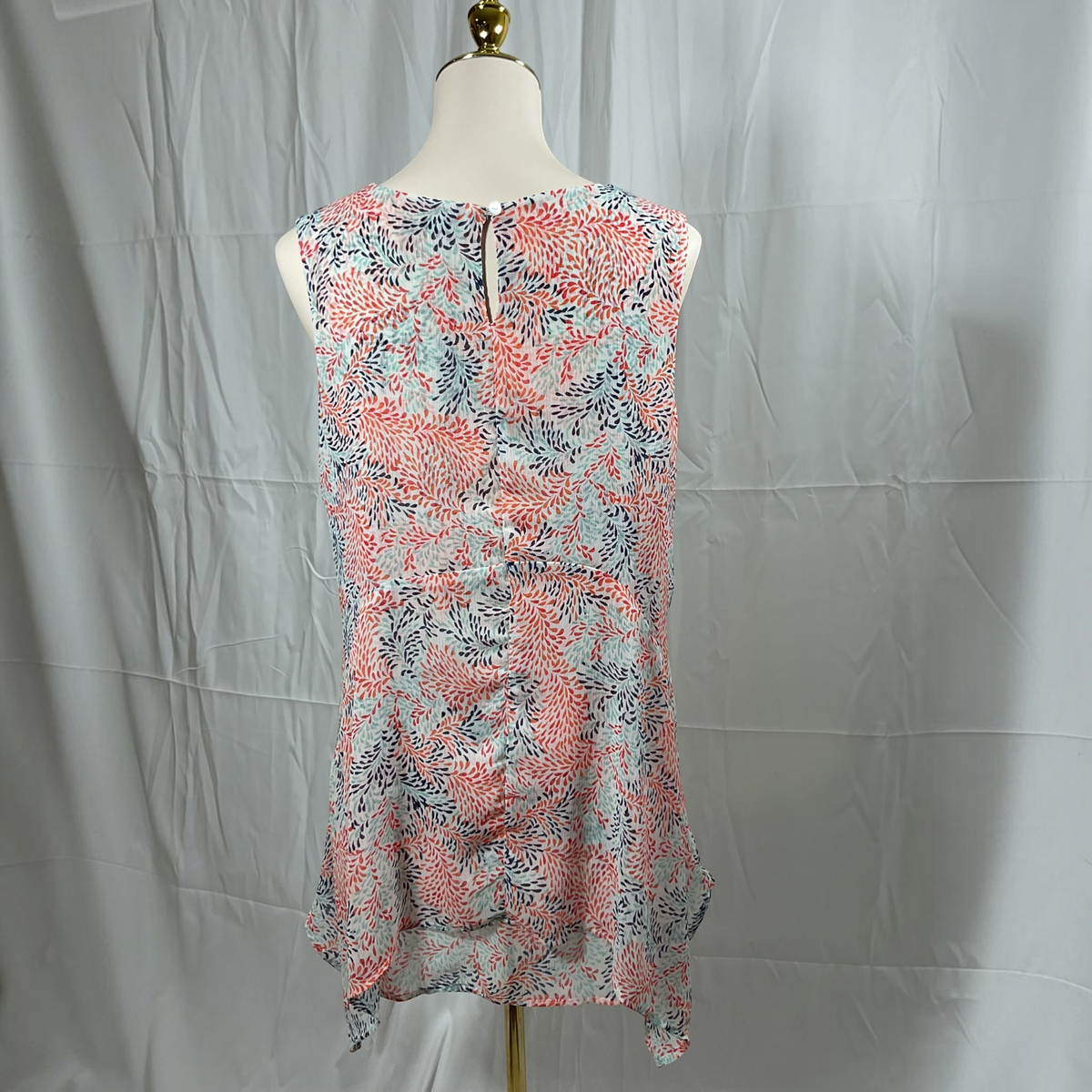 A sleeveless peplum top with a coral pink, light blue, and gray floral pattern, displayed on a cream mannequin torso against a wrinkled gray backdrop.