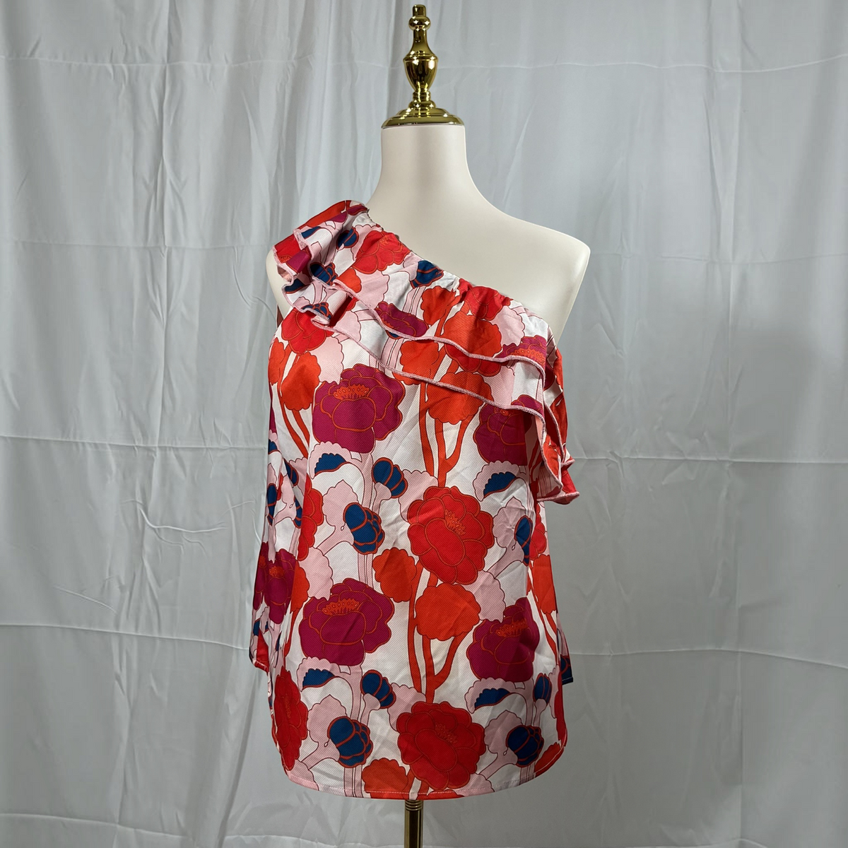 A one-shoulder blouse with a bold floral print, displayed on a cream mannequin torso with a gold neck cap against a wrinkled light gray fabric backdrop.
