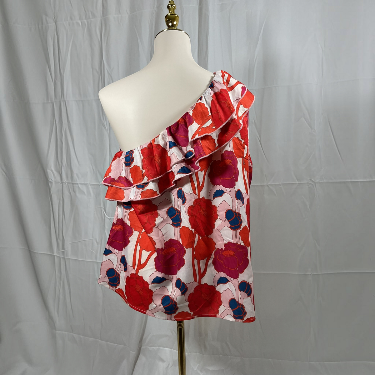 A one-shoulder ruffled top with a bold floral print in red, magenta, and navy blue on a white base, displayed on a cream mannequin torso against a wrinkled light gray backdrop.