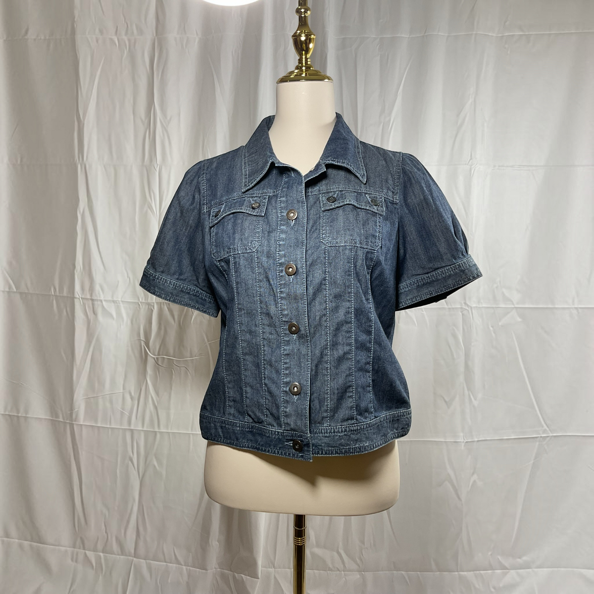A short-sleeve blue denim jacket with a button-down front and chest pockets, displayed on a cream mannequin torso against a wrinkled white fabric backdrop.