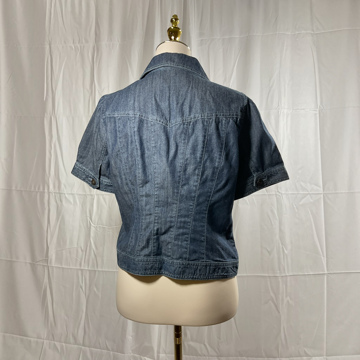 A short-sleeve denim jacket shown from the back on a cream dress form mannequin, set against a white wrinkled fabric backdrop.