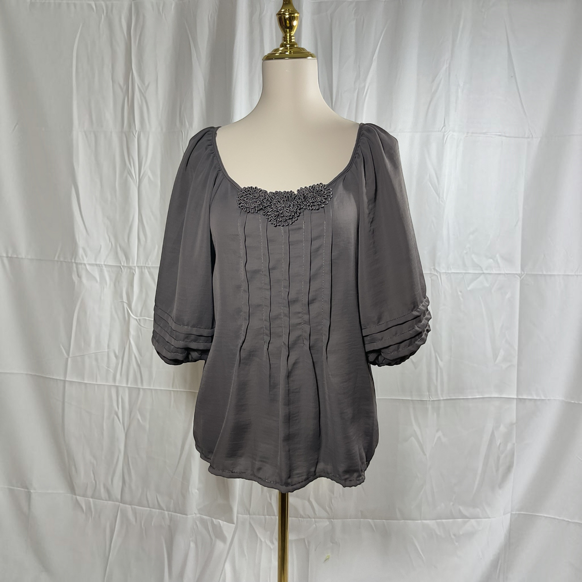 A gray short-sleeve blouse with a square neckline, gathered shoulders, and a decorative floral applique across the chest, displayed on a mannequin torso against a white fabric backdrop.