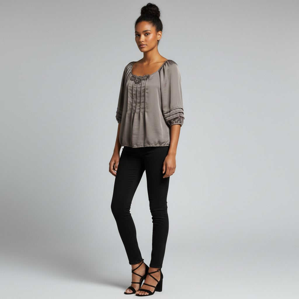 A woman modeling a taupe-gray short-sleeve blouse with gathered detailing and black skinny jeans against a light gray studio background.