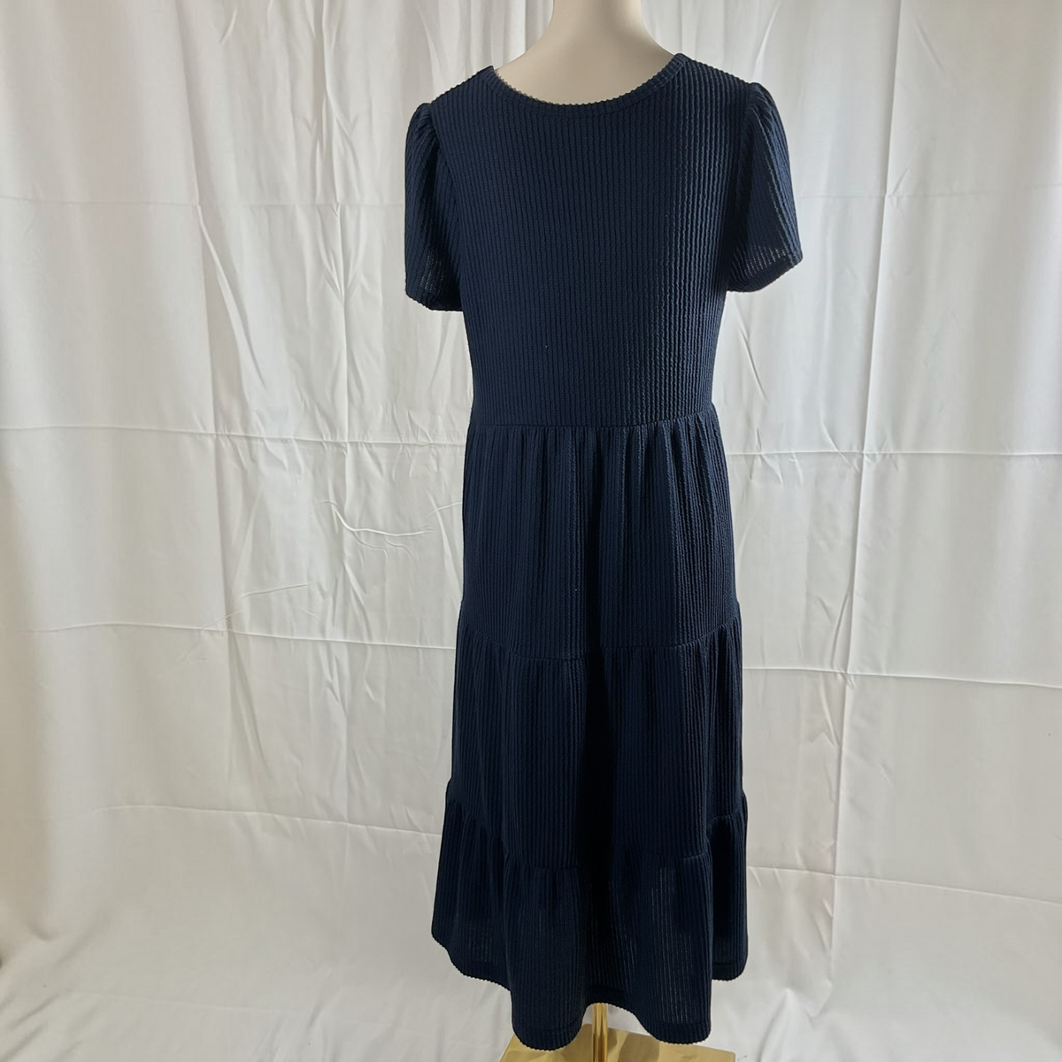 A dark navy blue ribbed knit dress with short sleeves and a tiered skirt, displayed on a mannequin against a wrinkled white backdrop.