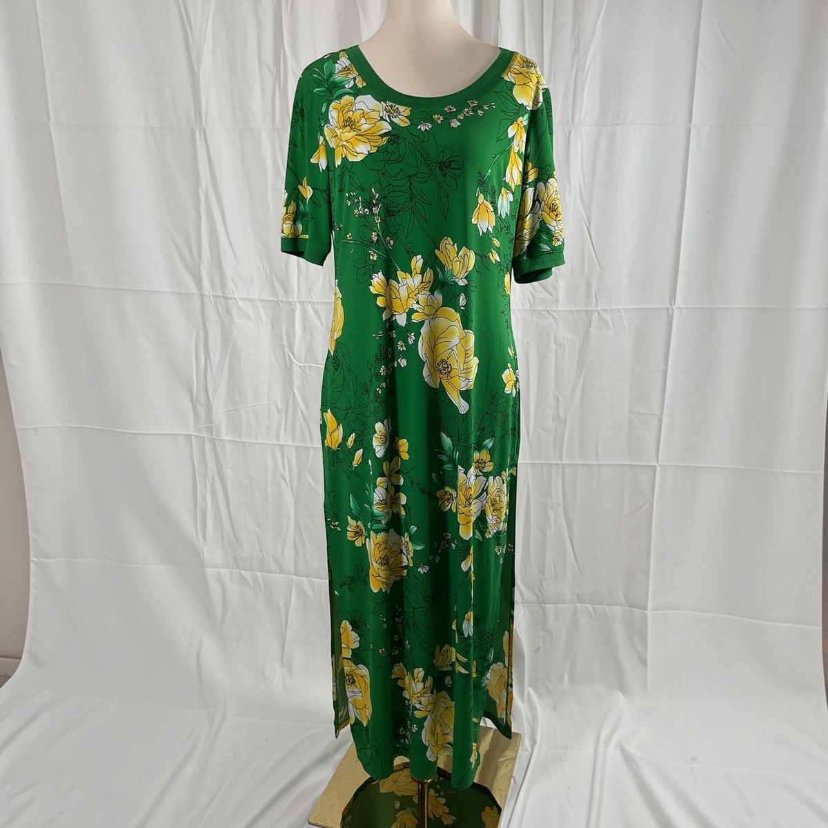 A green short-sleeve maxi dress with a yellow floral print, displayed on a mannequin against a white fabric backdrop.