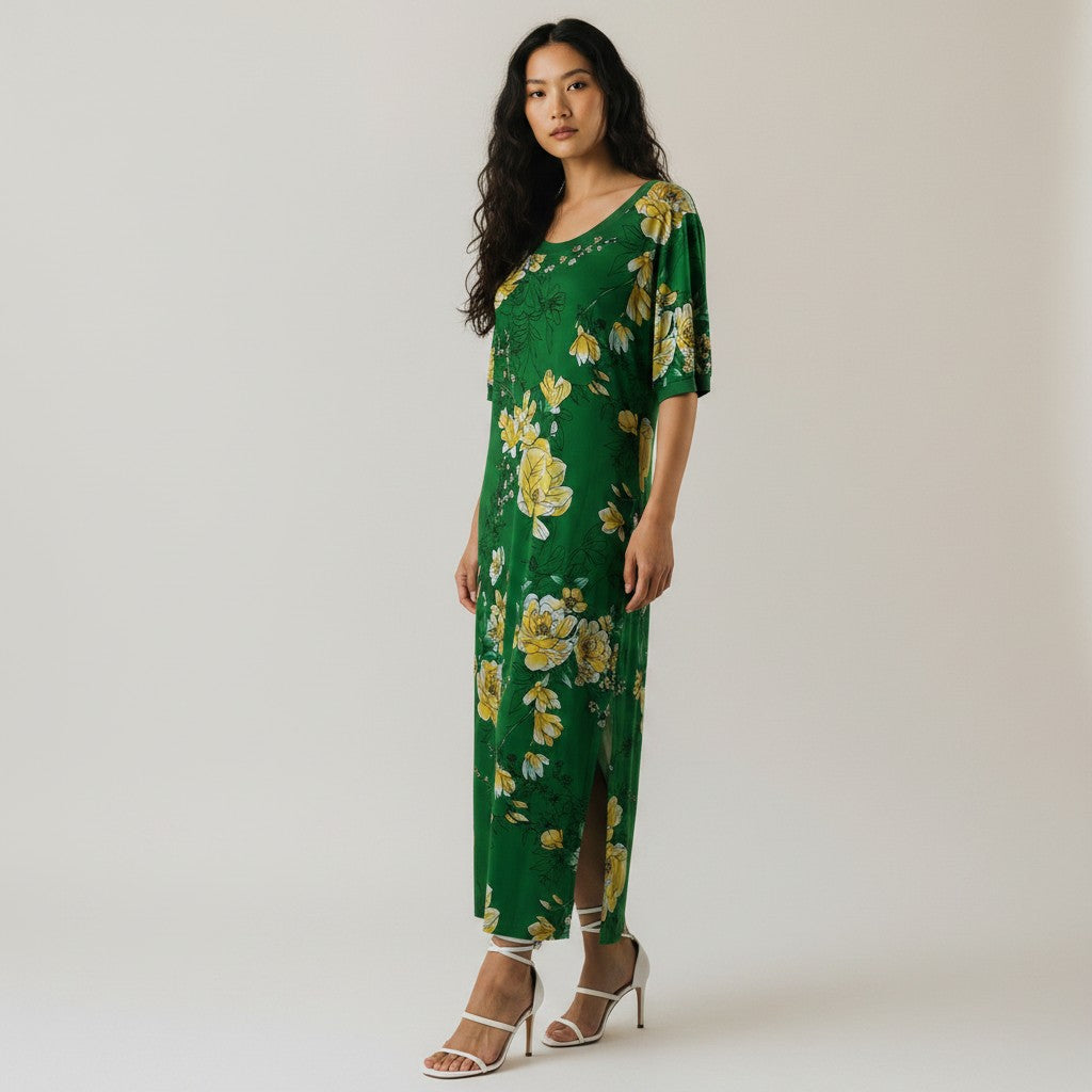 A woman modeling a green maxi dress with a yellow and white floral print, featuring a round neckline, short sleeves with lace detailing, and a side slit, paired with white strappy high-heeled sandals against a plain light background.