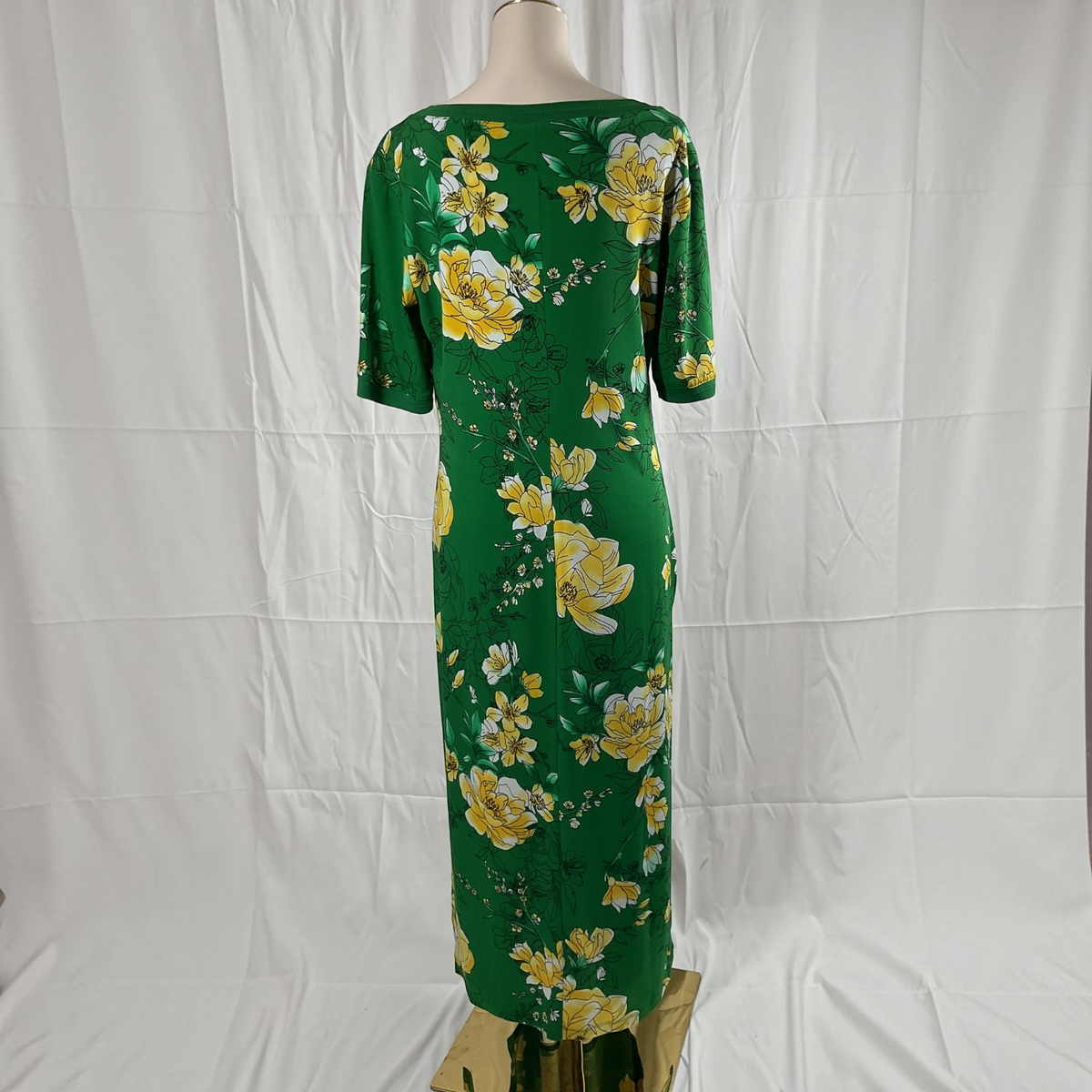 A green short-sleeve maxi dress with a yellow floral print, displayed on a mannequin against a white fabric backdrop.