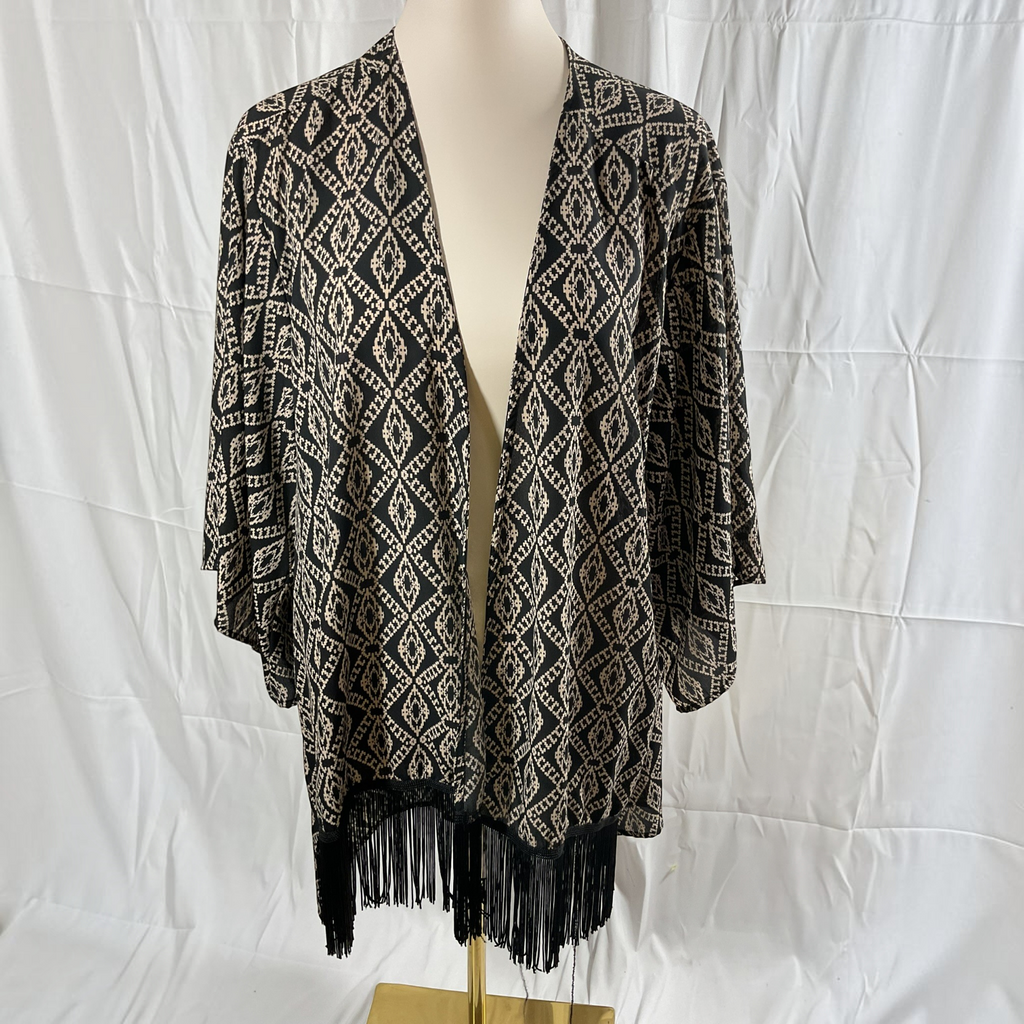 A black and beige geometric patterned open-front cardigan with black fringe along the hem, displayed on a cream mannequin torso against a white wrinkled fabric backdrop.