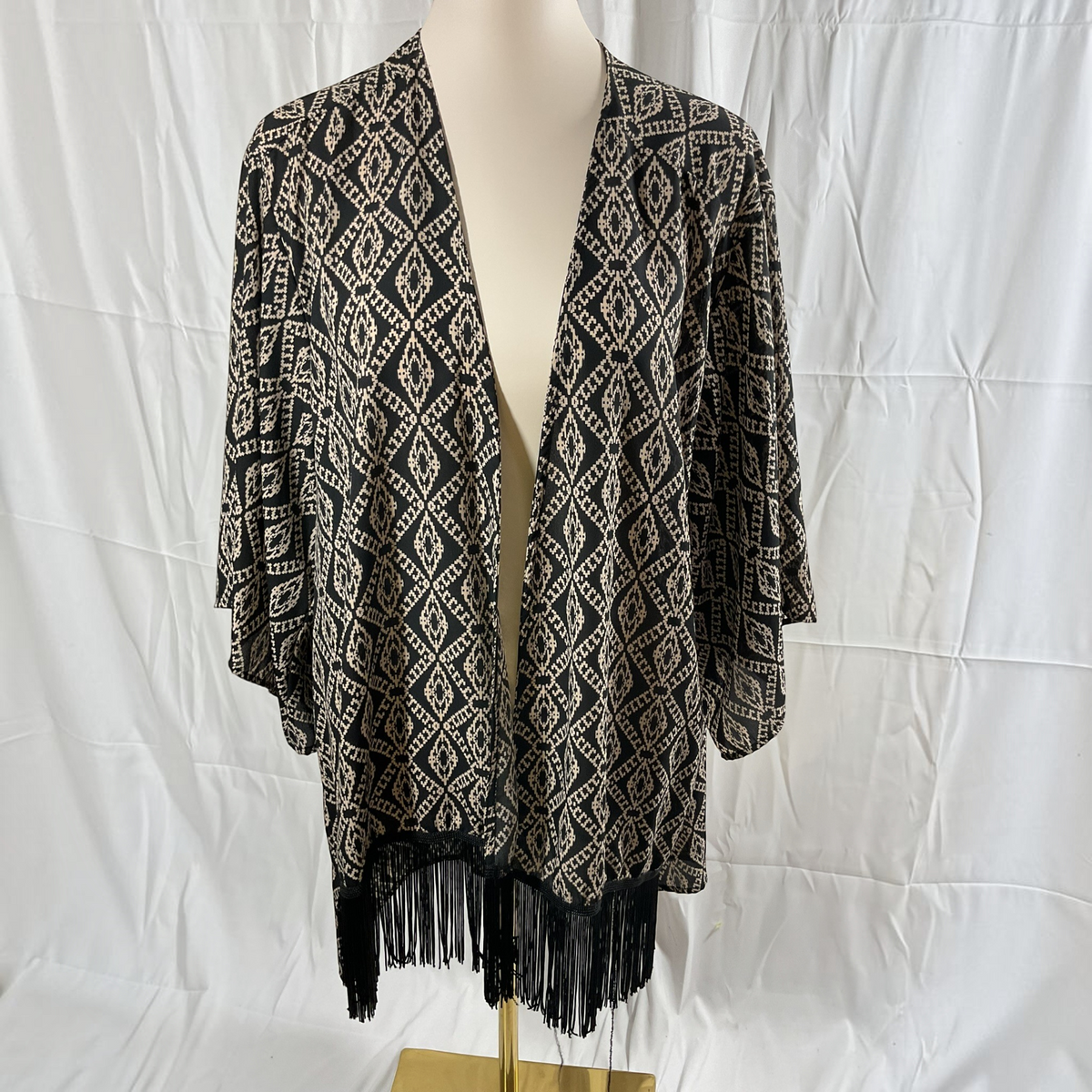 A black and beige geometric patterned open-front cardigan with black fringe along the hem, displayed on a cream mannequin torso against a white wrinkled fabric backdrop.