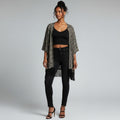 A woman stands against a light gray studio background wearing a black and beige patterned open-front fringed cardigan over a black crop top and black skinny jeans.