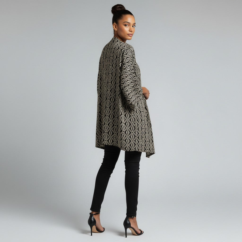 A woman modeling a long-sleeved, knee-length tunic with a black and beige geometric diamond pattern, paired with black skinny pants and black high-heeled sandals, photographed against a light gray studio background.