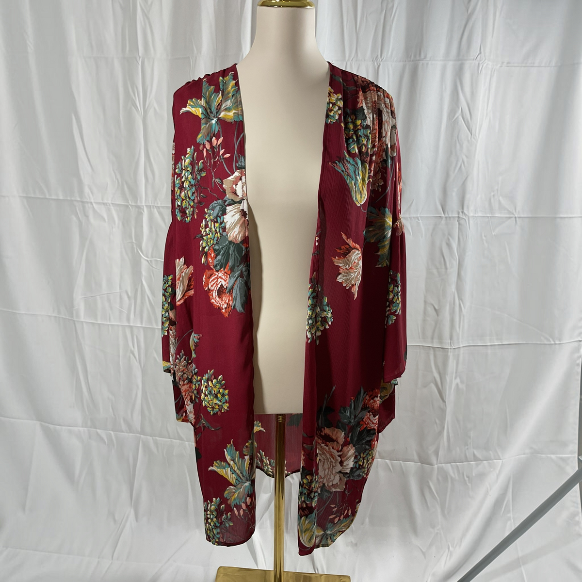 A burgundy open-front kimono cardigan with a colorful floral and botanical print, displayed on a cream mannequin torso with a gold stand against a white wrinkled fabric backdrop.