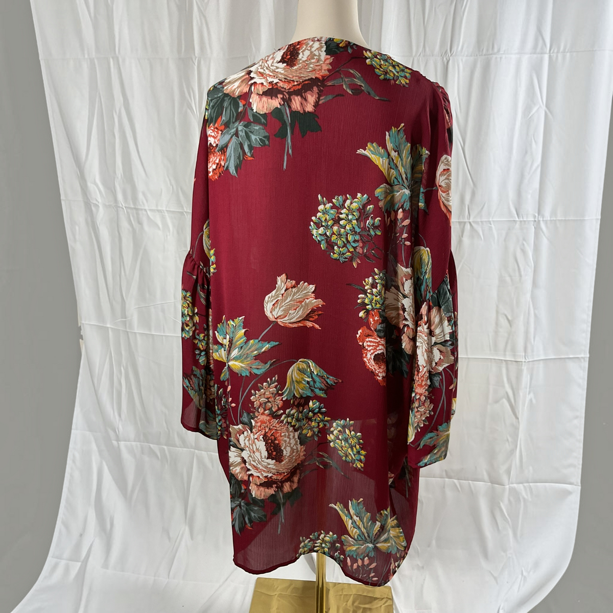 A long-sleeve floral blouse displayed on a dress form mannequin, featuring a deep burgundy base with large pink, peach, and green floral prints. The blouse is photographed against a white fabric backdrop.