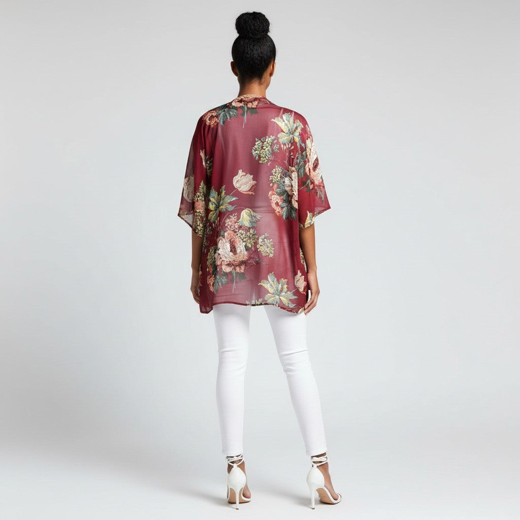 A woman viewed from behind wearing a sheer burgundy floral kimono top, white skinny pants, and white high heels against a light gray studio background.
