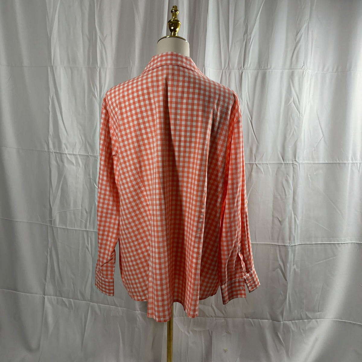 A long-sleeve gingham check shirt in orange and white, displayed on a dress form mannequin against a wrinkled white fabric backdrop.