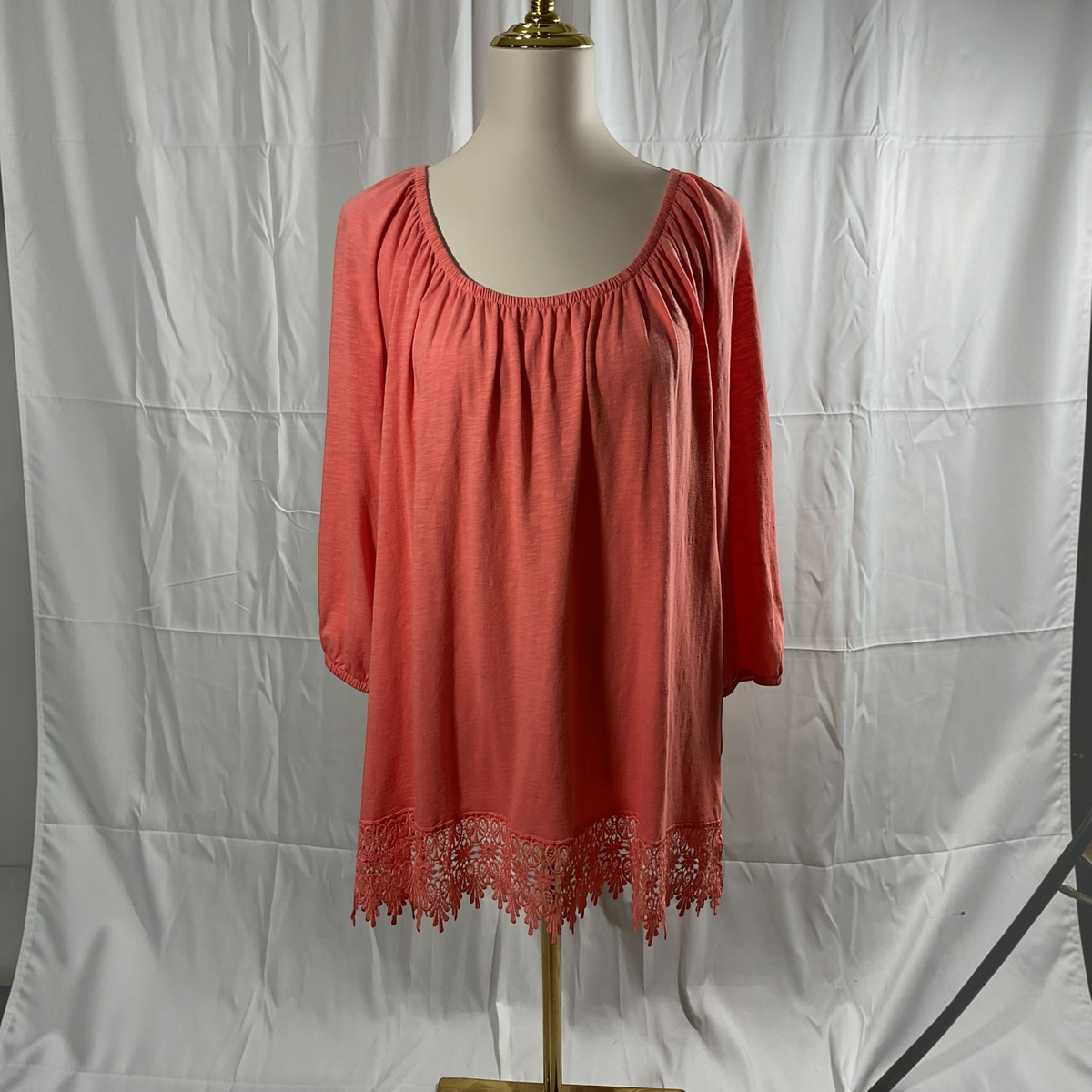 A coral pink long-sleeve top with a gathered neckline and lace hem, displayed on a mannequin torso against a wrinkled white studio backdrop.