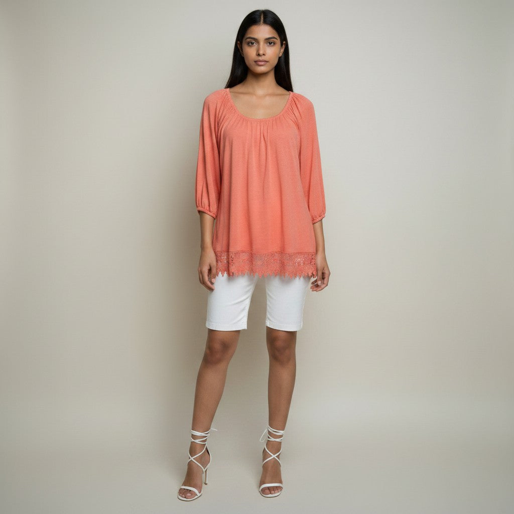 A woman modeling a loose, short-sleeve peach top with a gathered neckline and lace hem, paired with white shorts and white strappy high-heeled sandals, against a plain light beige studio background.