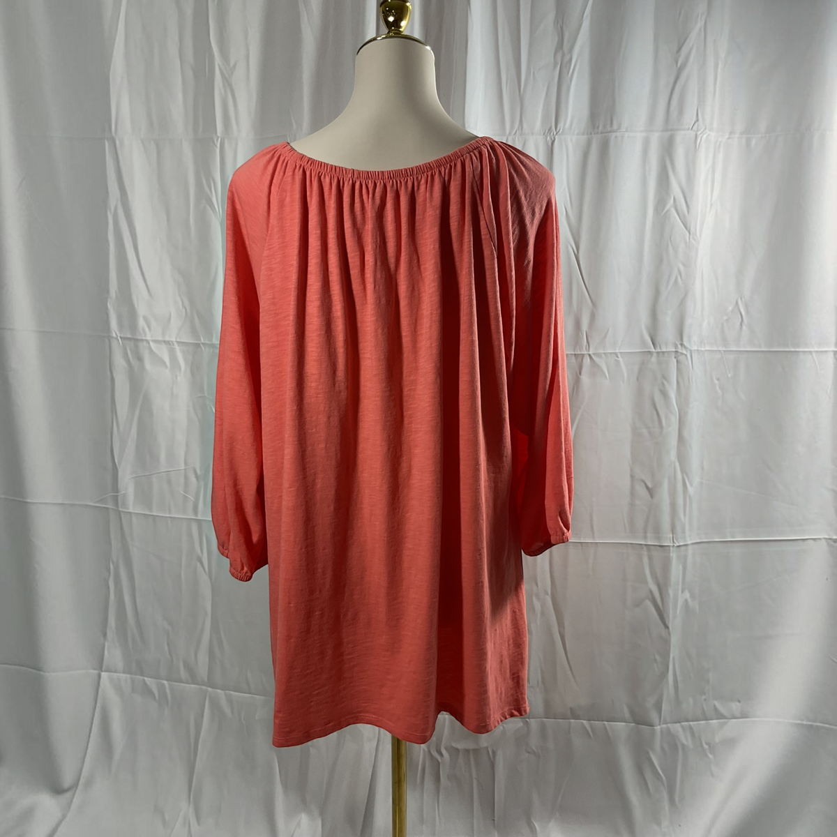A coral pink long-sleeve top with a gathered, elasticized neckline, displayed on a cream dress form mannequin against a wrinkled white fabric backdrop.