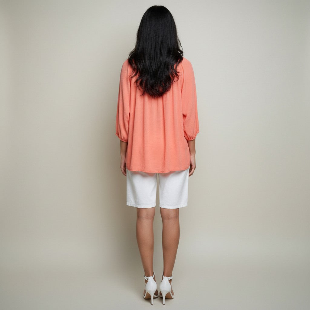A woman viewed from behind wearing a peach-colored, three-quarter sleeve blouse and white shorts against a plain light beige studio background.