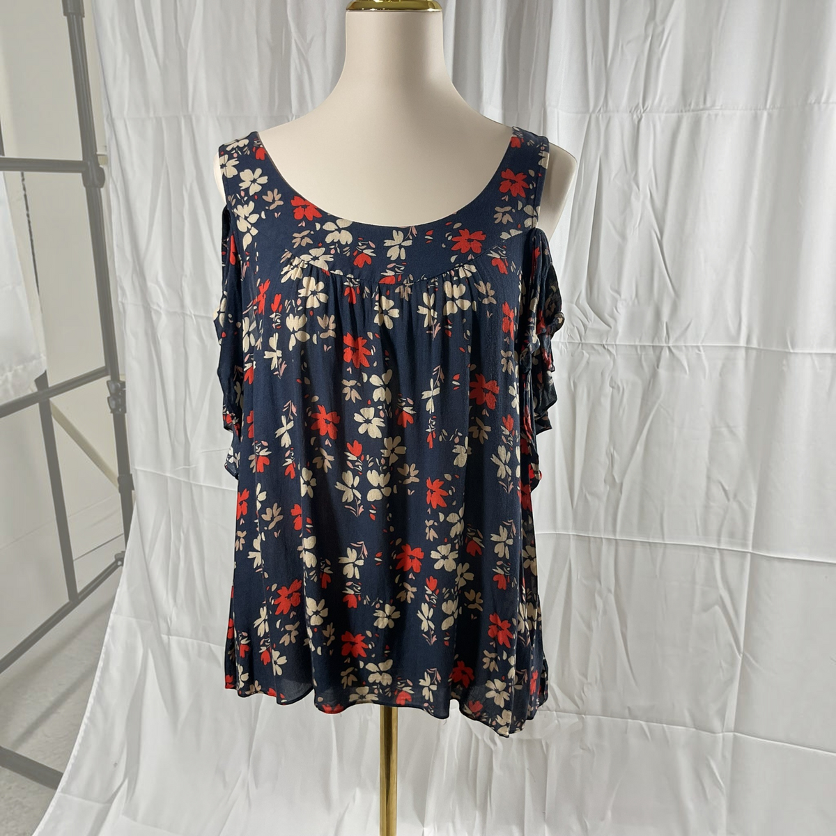 A sleeveless floral top displayed on a mannequin torso against a white draped backdrop. The top has a dark navy base with a red and cream floral print, and ruffled side details.