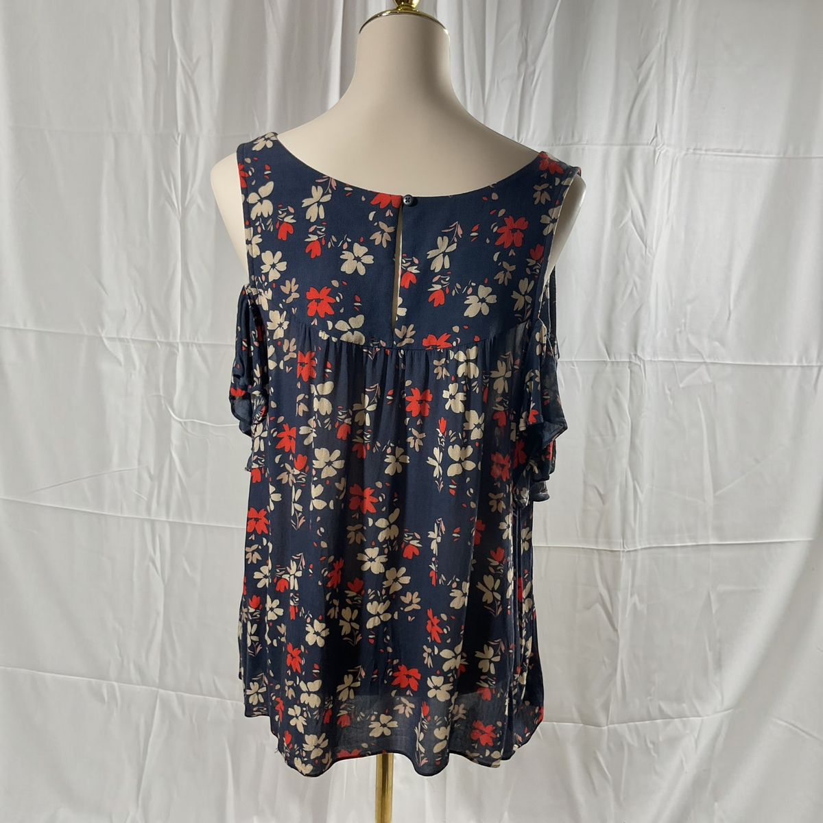 A sleeveless navy blue blouse with a red and white floral pattern, displayed on a cream mannequin torso against a wrinkled white fabric backdrop.