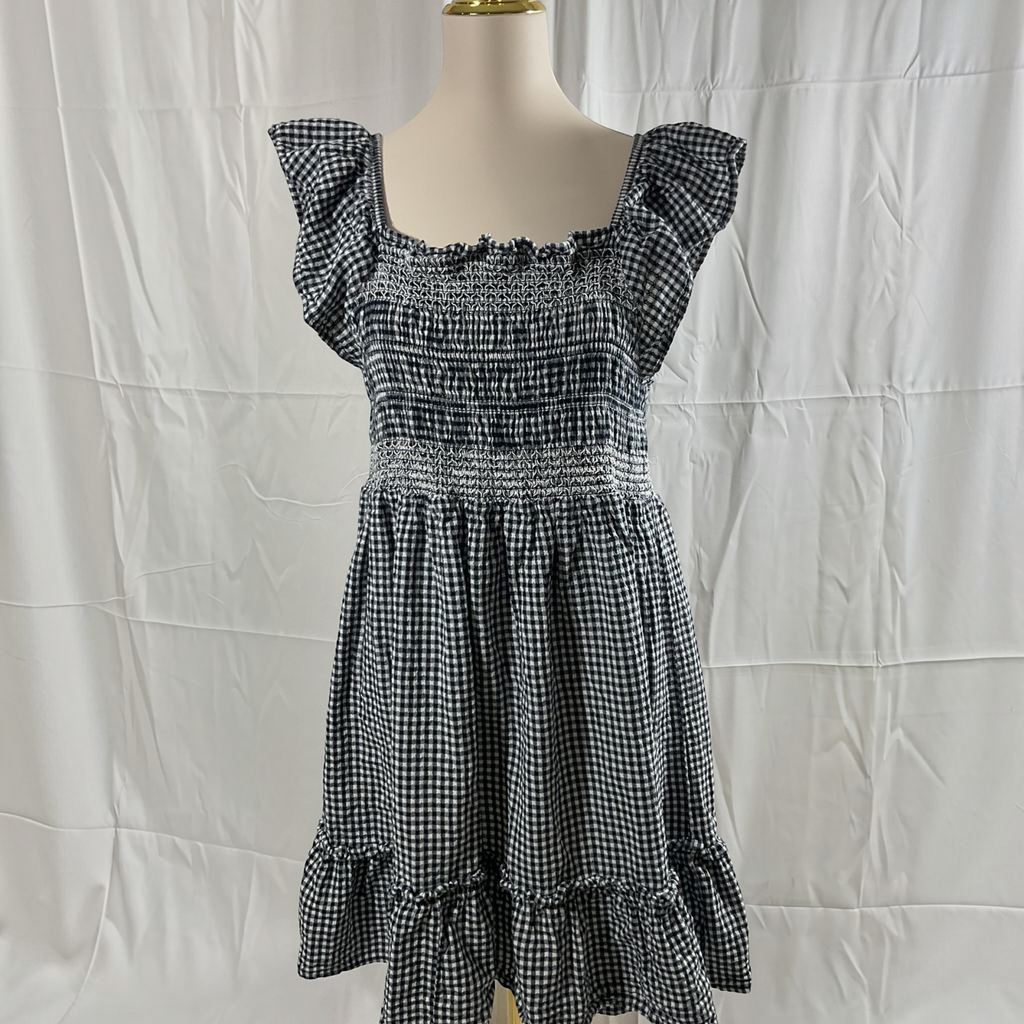 A black and white gingham dress displayed on a mannequin torso against a wrinkled white fabric backdrop. The dress features ruffled cap sleeves, a smocked bodice, and a tiered skirt.