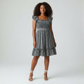 A woman wearing A black and white gingham dress displayed on a mannequin torso against a wrinkled white fabric backdrop. The dress features ruffled cap sleeves, a smocked bodice, and a tiered skirt.