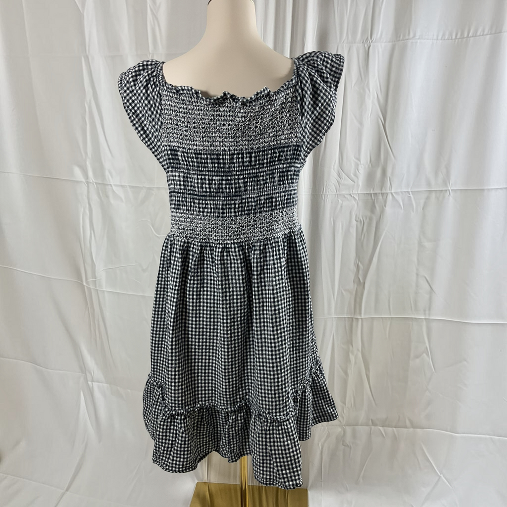 A black and white gingham dress displayed on a mannequin torso against a white fabric backdrop. The dress features a smocked bodice, cap sleeves, and a tiered ruffle hem.