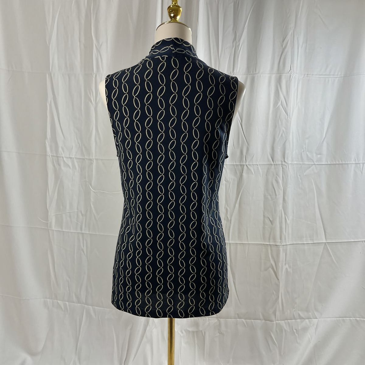 A sleeveless collared top or tunic displayed on a dress form mannequin, shown from the back. The garment has a dark navy base with a repeating vertical pattern of thin white wavy lines, set against a wrinkled white fabric backdrop.