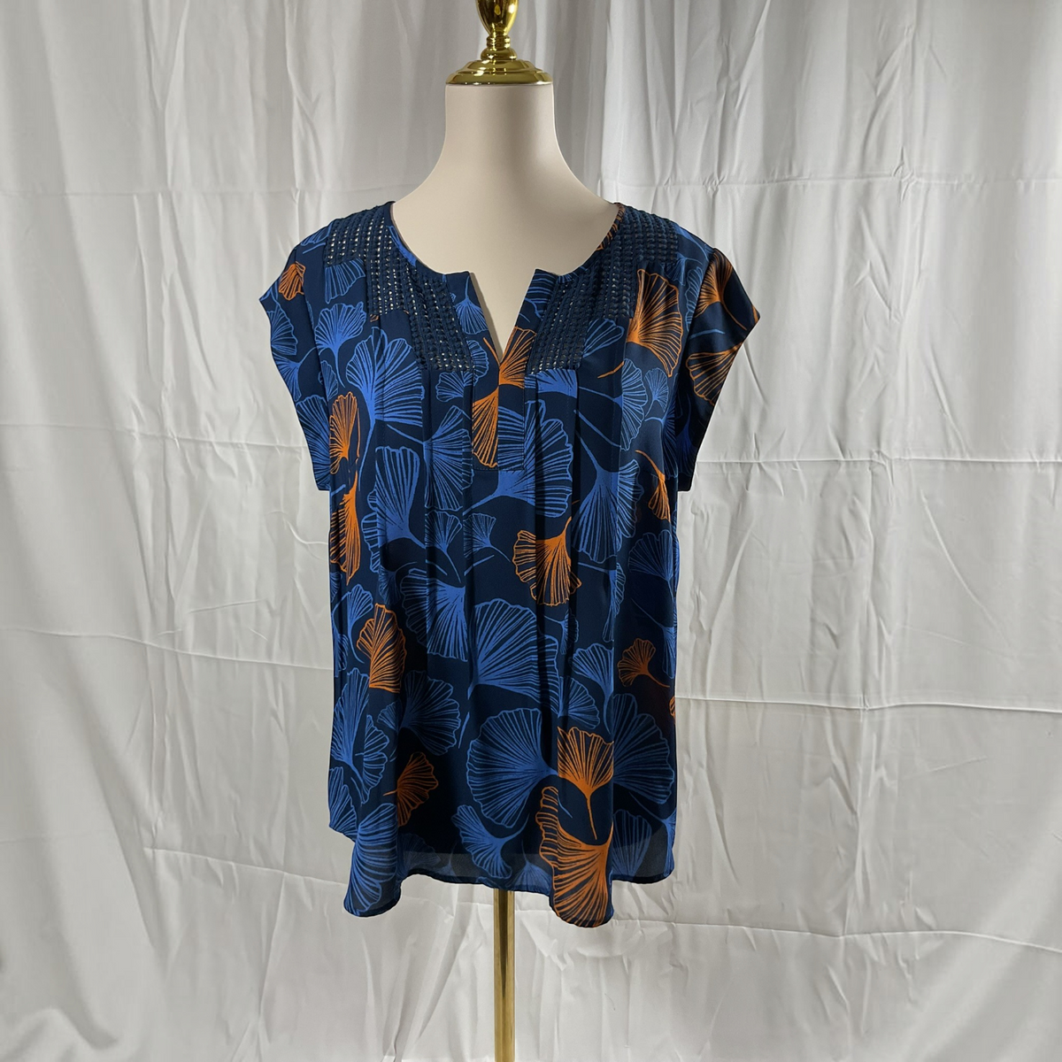 A short-sleeve blouse with a split neckline, displayed on a mannequin torso. The top features a blue and orange ginkgo leaf pattern on a dark navy base, set against a wrinkled white fabric backdrop.
