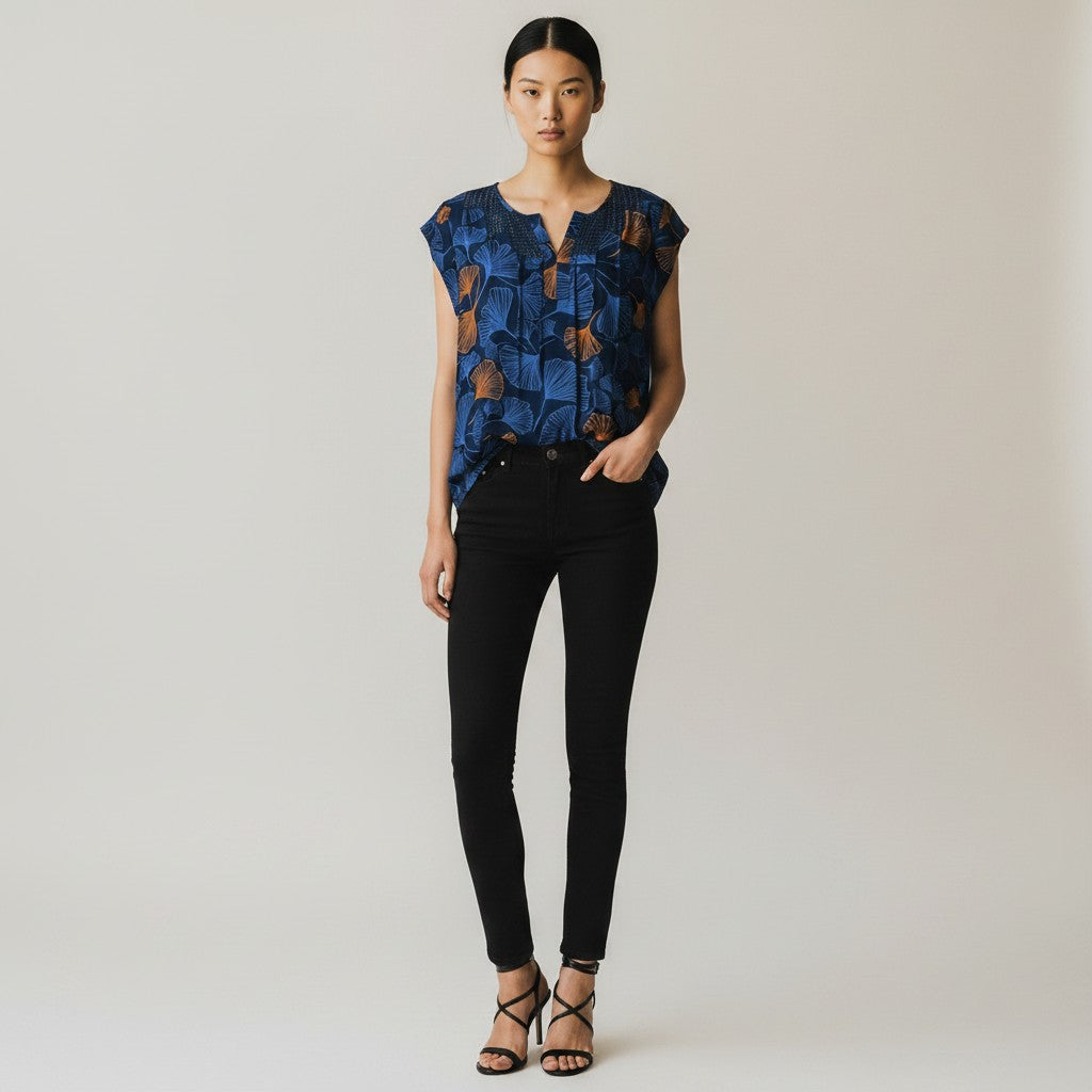 A woman modeling a short-sleeve V-neck blouse with a blue and brown abstract print, paired with black skinny pants and black strappy heels, against a light gray studio background.