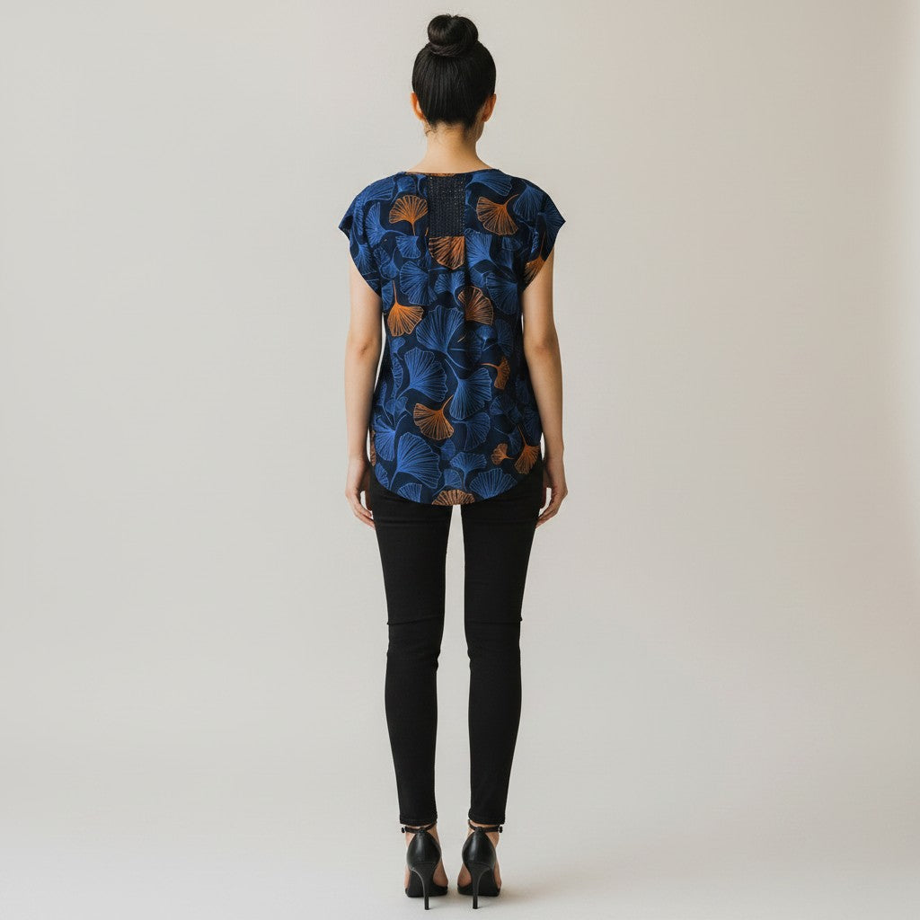 A woman viewed from behind wearing a short-sleeve blue top with a brown ginkgo leaf pattern, paired with black skinny pants and black high heels, against a light gray studio background.