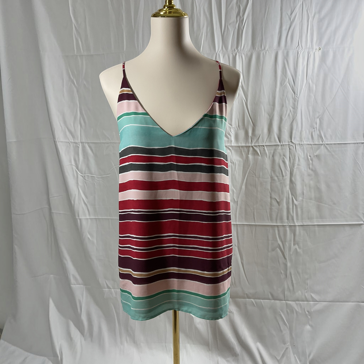 A sleeveless striped camisole tank top displayed on a cream mannequin torso against a wrinkled white fabric backdrop. The top features horizontal stripes in teal, burgundy, dark purple, pink, and thin beige lines.