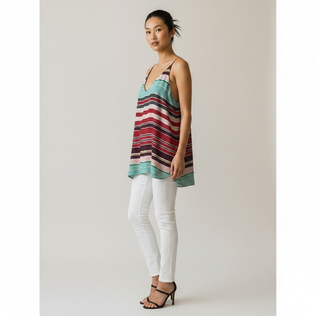 A woman modeling a sleeveless striped tunic top with horizontal bands of teal, red, black, and white, paired with white pants and black sandals, against a light gray studio background.