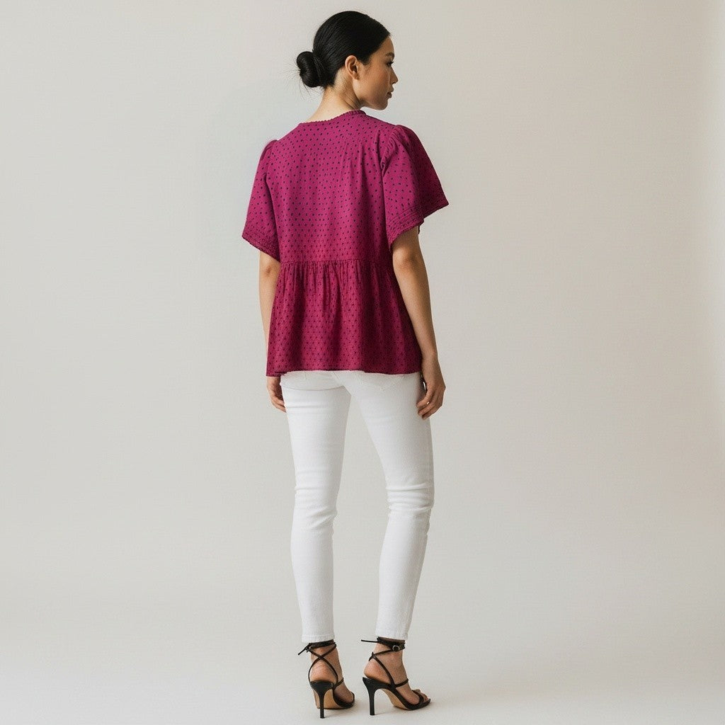 A woman is shown from the back wearing a short-sleeved magenta top with a small white geometric pattern, paired with white skinny pants and black high-heeled sandals, against a plain light gray background.