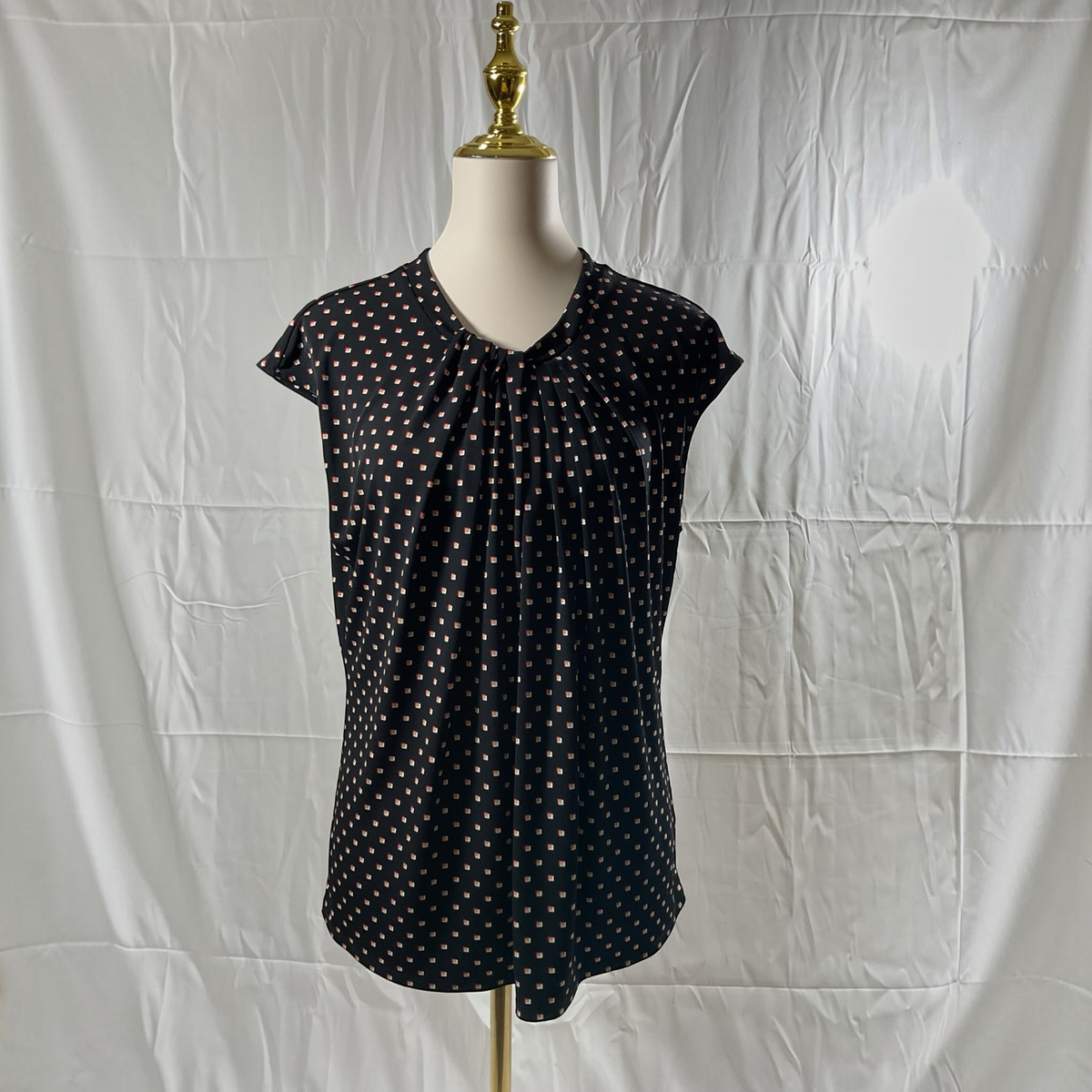 A black short-sleeve blouse with a white polka dot pattern, displayed on a cream mannequin torso with a gold neck cap against a wrinkled white fabric backdrop.