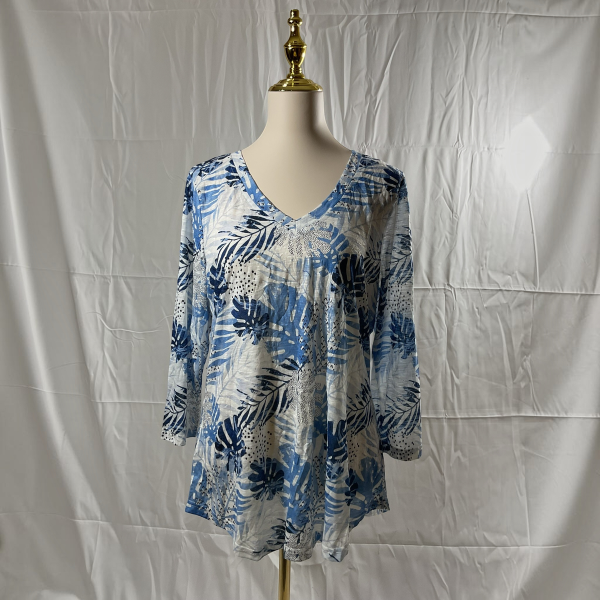 A women's long-sleeve V-neck tunic top with a blue, navy, and white tropical leaf pattern, displayed on a cream mannequin torso with a gold finial against a wrinkled white fabric backdrop.