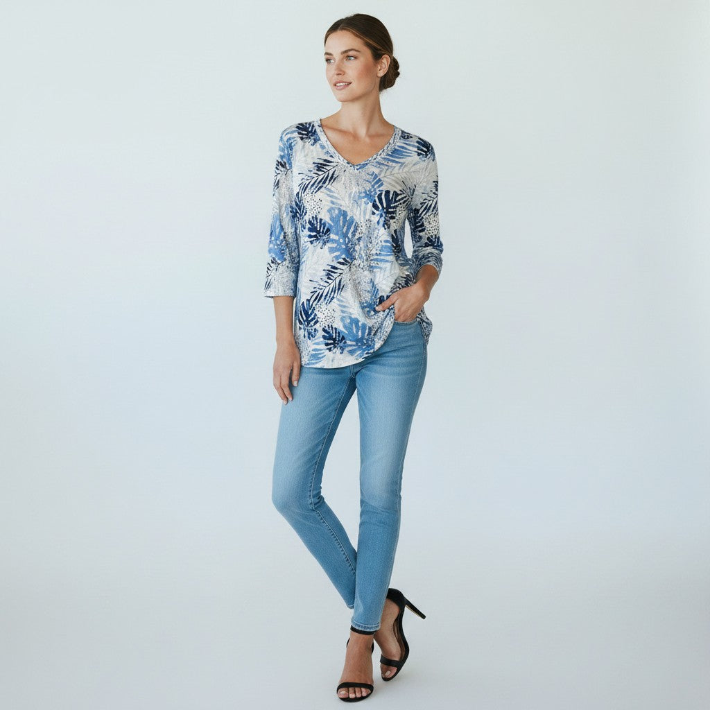 A woman modeling a blue and white tropical leaf print blouse with three-quarter sleeves, paired with light-wash blue jeans and black high-heeled sandals, against a light gray studio background.
