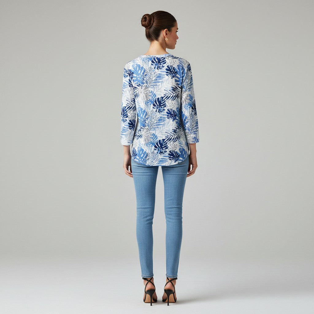 A woman viewed from the back wearing a white long-sleeve blouse with a blue tropical leaf pattern, paired with light blue skinny jeans and black high-heeled sandals, against a neutral gray studio background.