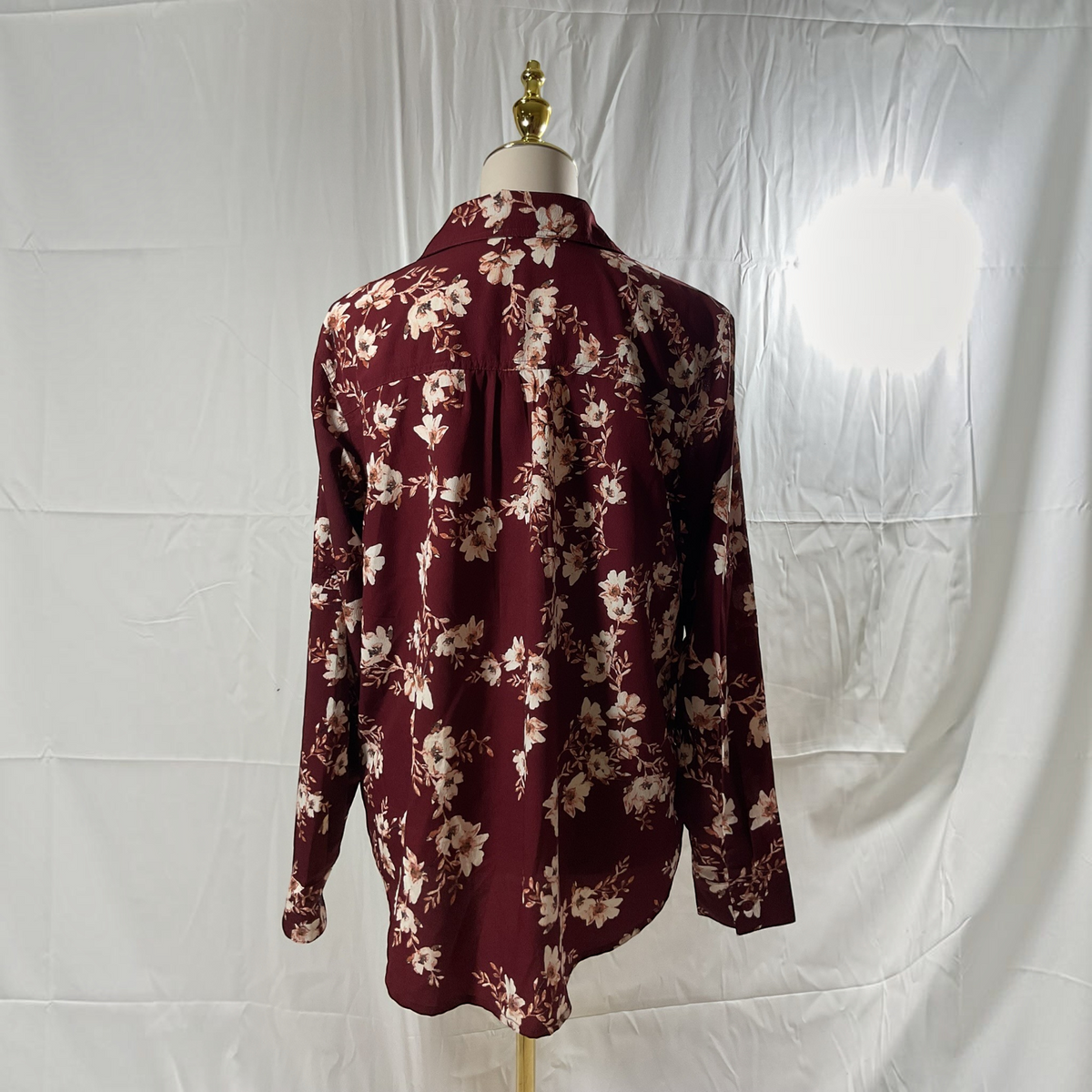 A long-sleeve button-up blouse with a floral print, displayed on a dress form against a wrinkled white fabric backdrop.