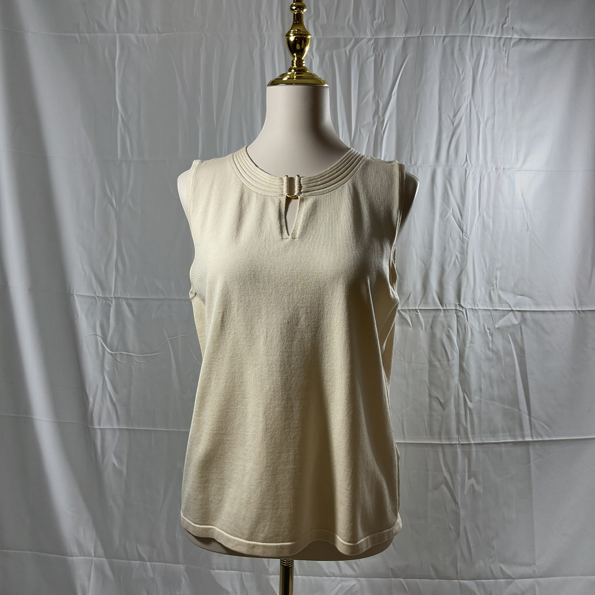 A sleeveless cream-colored knit top displayed on a dress form mannequin against a wrinkled white fabric backdrop.