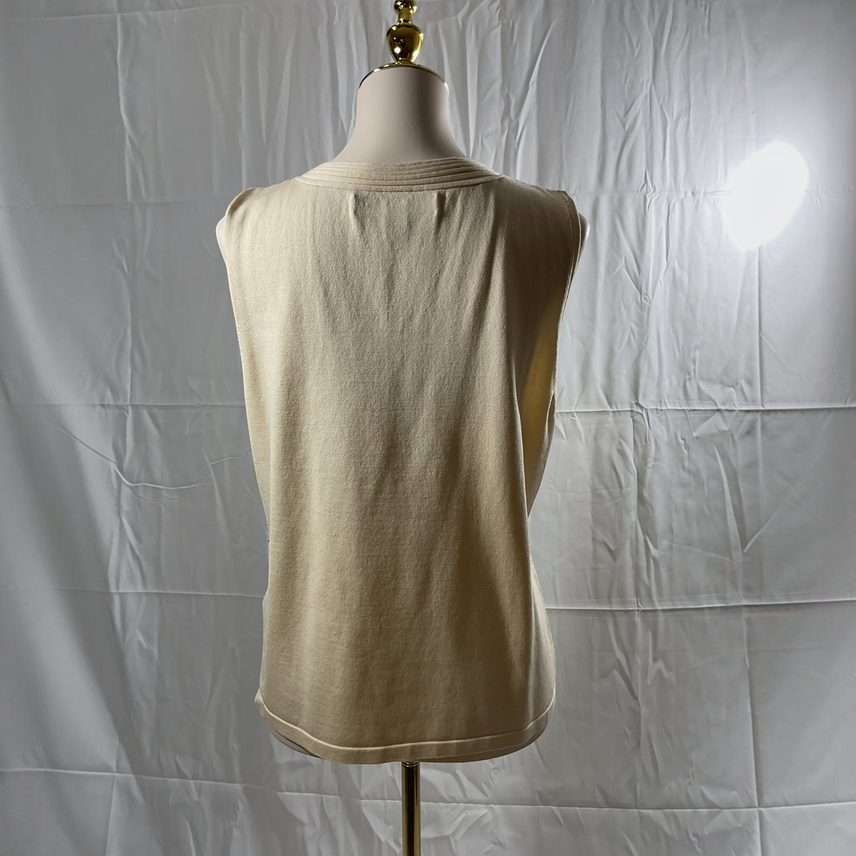 A sleeveless knit top in a light cream or beige color, displayed on a dress form mannequin from the back against a wrinkled white fabric backdrop.