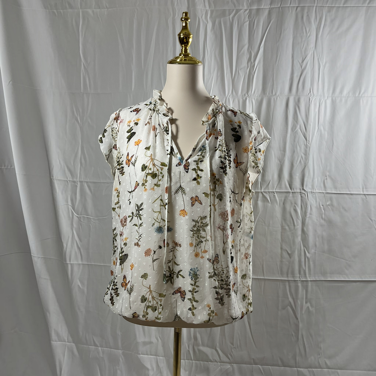 A white short-sleeve blouse with a colorful floral and butterfly print, displayed on a dress form mannequin against a wrinkled white backdrop.